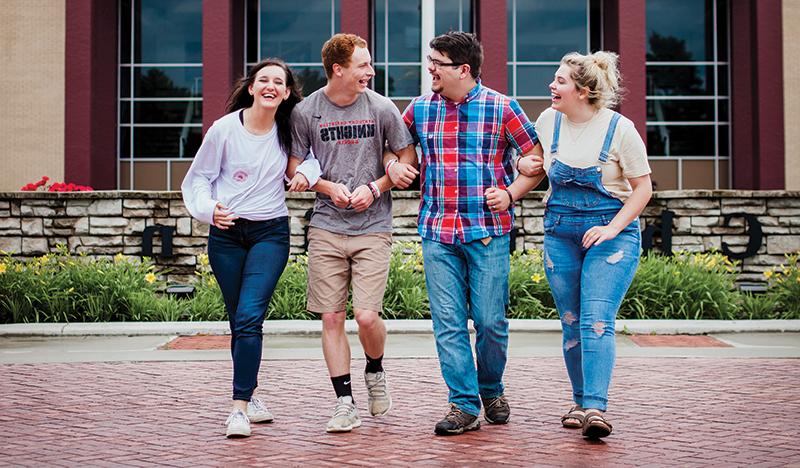 Four people walking and laughing together outside, with a brick building in the background. Two on the left wear casual outfits, and two on the right wear t-shirts and jeans.